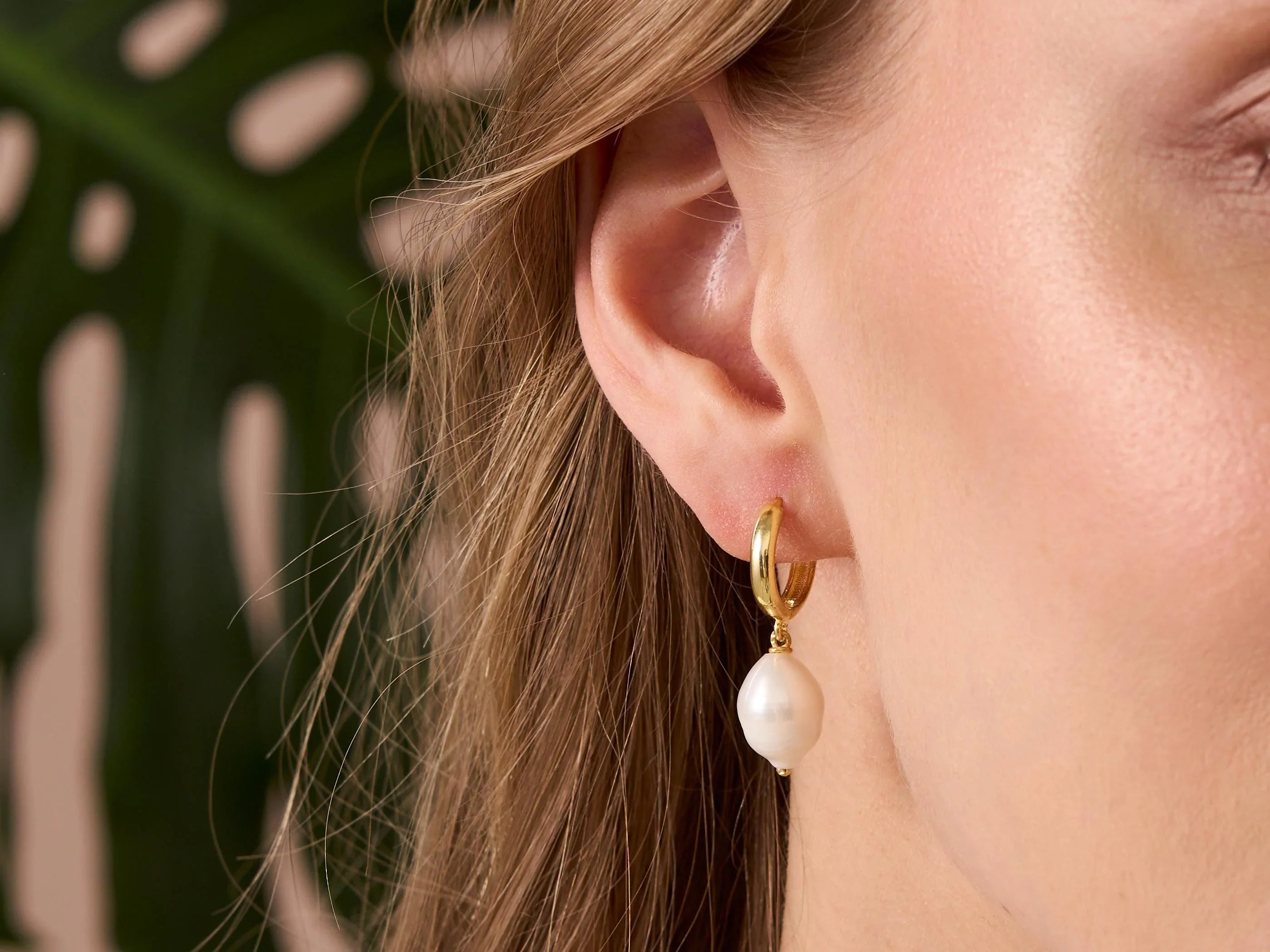 Close-up of a person wearing a gold hoop earring with a pearl drop against a blurred natural background.